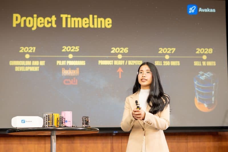 SooAh Lay presenting the Avakas Lab project timeline on stage, CanSats laid out on the table beside her.