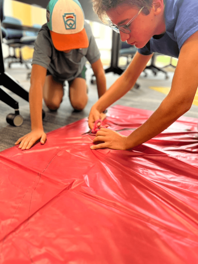 Two students kneeling on the floor of a classroom, cutting and shaping red parachute fabric for their CanSat recovery system.